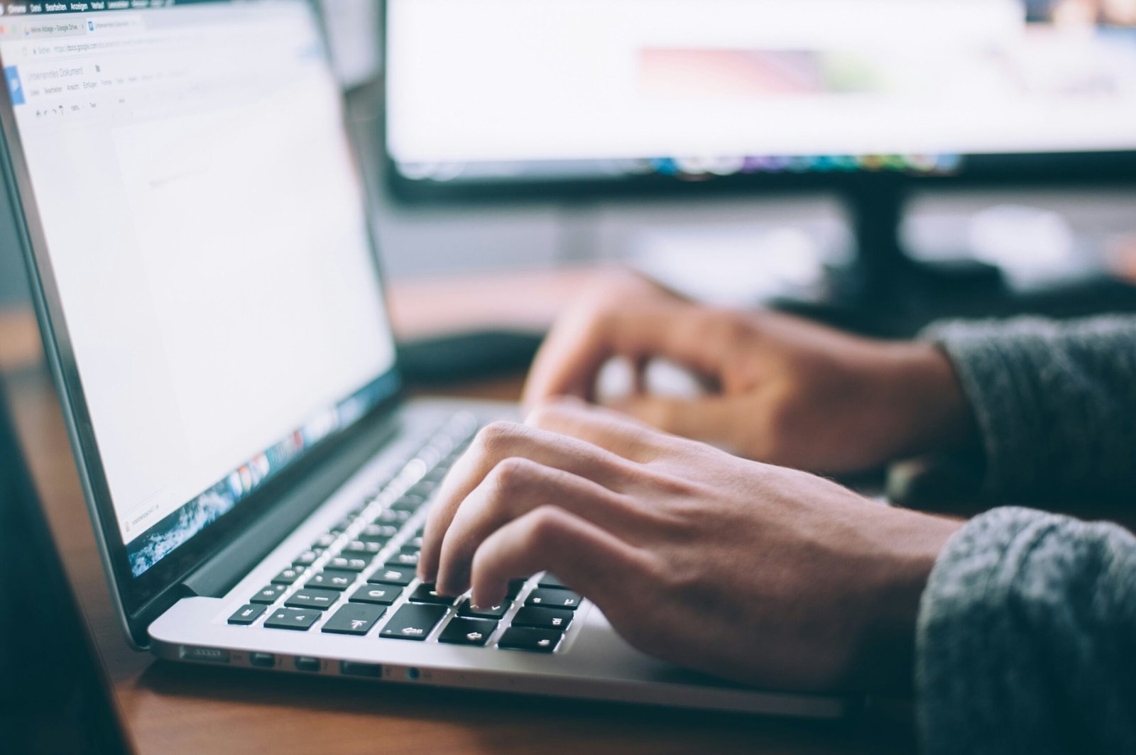 A close-up shot of a person's hands typing on the keyboard of a silver laptop, with a blurred computer monitor in the background.